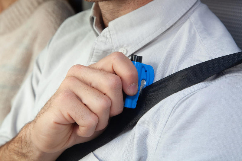 Image of a person's hand holding a blue Resqme tool against a black car seatbelt, demonstrating its use for cutting.