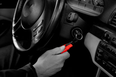 Image, mostly black and white, showing a hand holding a red Resqme tool attached to car keys in the ignition, with the car's steering wheel and dashboard visible.