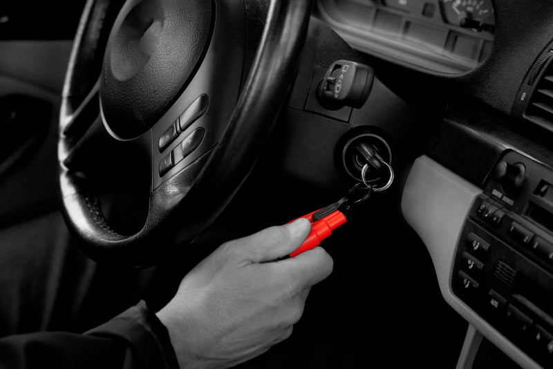 Image, mostly black and white, showing a hand holding a red Resqme tool attached to car keys in the ignition, with the car's steering wheel and dashboard visible.