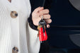 Image of a person's hand holding a set of keys with a red Resqme tool attached, against a blurry background of a car and a white knitted cardigan.