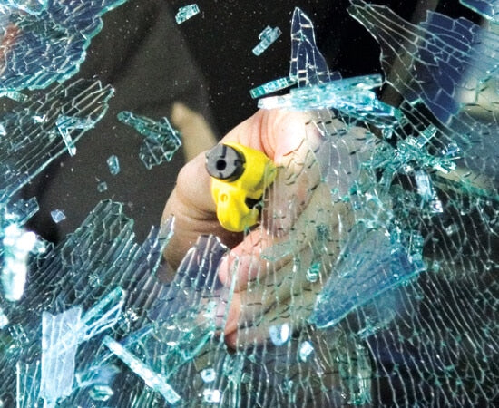 Image of a hand holding a yellow Resqme tool breaking through a car window, surrounded by shattered glass.