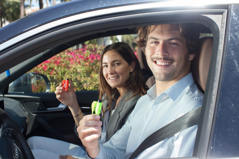 Image of a smiling man and woman in a car, both holding up Resqme keychain tools (the man holding a green one, the woman a red one), with flowers visible outside the car window.