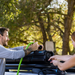 Image of a man and a woman setting up the RinseKit Portable Rack Shower on a vehicle's roof rack, with the man holding a green hose and the woman connecting a black cable, against a backdrop of green trees