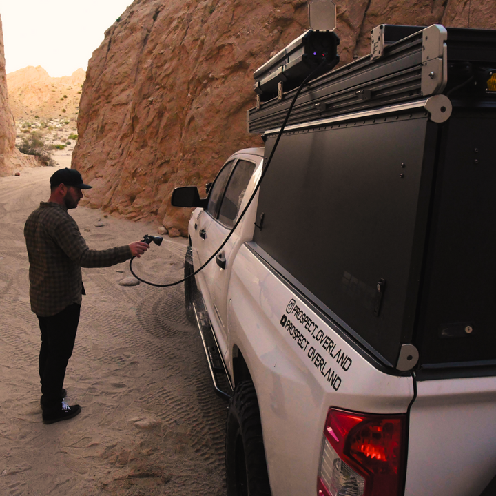 Image of a man in a cap using the RinseKit Portable Rack Shower to spray water, with the unit mounted on the roof rack of a white pickup truck, in a sandy canyon landscape, showing "PROSPECT OVERLAND" branding on the truck's rear