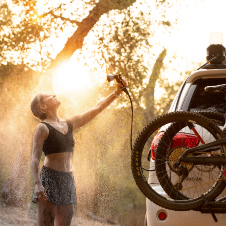 Image of a woman with a dirt-smudged face and body rinsing herself off with a spray of water from the RinseKit Portable Rack Shower, which is mounted to a vehicle with a bike rack and a mountain bike attached, with sunlight shining through the trees in the background