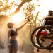 Image of a woman with a dirt-smudged face and body rinsing herself off with a spray of water from the RinseKit Portable Rack Shower, which is mounted to a vehicle with a bike rack and a mountain bike attached, with sunlight shining through the trees in the background
