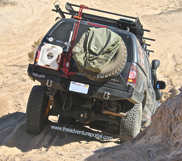 Image of off-road SUV driving on sandy terrain with rear-mounted RotopaX white water container, spare tire, and roof rack accessories, angled rear view in desert setting.