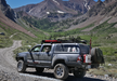 Image of off-road pickup truck with roof rack and rear-mounted RotopaX white water container parked on rocky mountain trail, side angle view in alpine landscape.