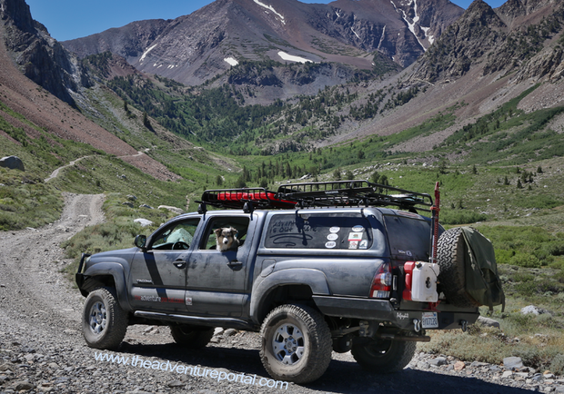 Image of off-road pickup truck with roof rack and rear-mounted RotopaX white water container parked on rocky mountain trail, side angle view in alpine landscape.