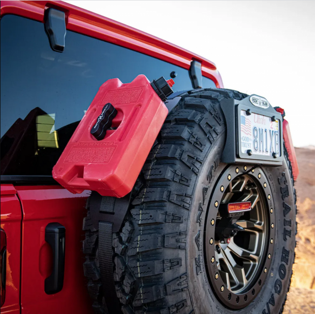 Image of RotopaX 1 Gallon Gasoline fuel container mounted on rear spare tire of red off-road vehicle, red can with black mount secured by strap, angled side view outdoors.