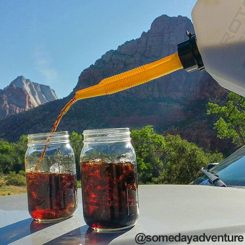 Image of RotopaX 1 Gallon Water container pouring liquid through yellow spout into glass jars, outdoor mountain landscape background with vehicle hood in foreground.