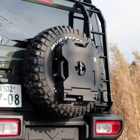 Black RotoPaX fuel can mounted on a spare tire of a green SUV, with a ladder attached to the side, angled rear view.