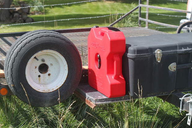 Image of a red RotopaX fuel container mounted on a utility trailer beside a spare tire and black storage box, outdoor side view showing secure transport setup.