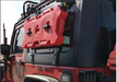 Red RotoPaX gasoline can with two black RotoPaX mounts, attached to the side window of a red Jeep, angled side view.