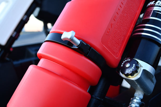 Close-up of a red Rotopax 1.5-gallon fuel can, mounted with a black clamp and silver T-handle bolt next to an adjustable shock absorber on an off-road vehicle.