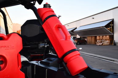 Red 1.5-gallon fuel can by Rotopax mounted to the roll cage of an off-road vehicle, with a large warehouse and mountainous landscape in the background.