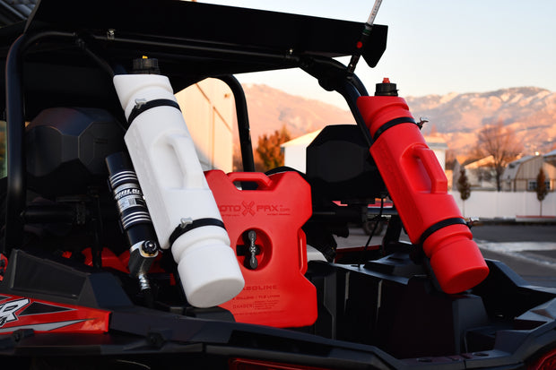 Three Rotopax fuel containers—one white 1.5-gallon, one red 2-gallon, and one red 1.5-gallon—are mounted on the back of an off-road vehicle with a scenic mountain range in the background.