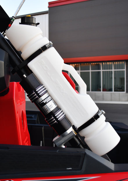 A white 1.5-gallon Rotopax fuel can is mounted with black clamps to the roll cage of an off-road vehicle, positioned next to a black shock absorber, with a modern building in the background.