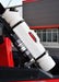 A white 1.5-gallon Rotopax fuel can is mounted with black clamps to the roll cage of an off-road vehicle, positioned next to a black shock absorber, with a modern building in the background.