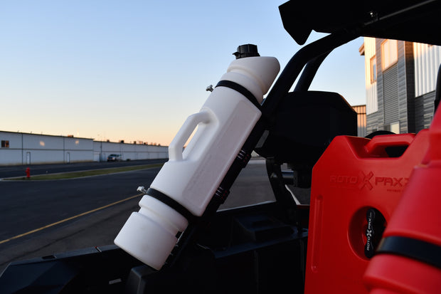 A white 1.5-gallon Rotopax fuel can is mounted on the roll cage of an off-road vehicle, with part of a red Rotopax container visible and an airport tarmac at sunset in the background.