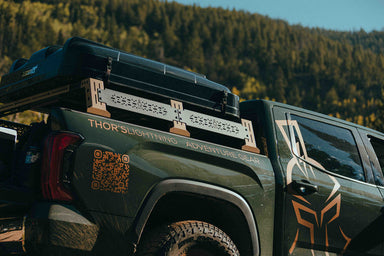 Image of Thors Lightning truck bed rack in midnight black mounted on pickup with rooftop tent, angled rear side view in outdoor setting.