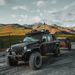Image of a black Jeep Gladiator overland truck with an extensive roof rack and an antenna, towing a small orange trailer on a dirt road. The vehicle is set against a backdrop of rolling mountains with patches of green and yellow vegetation under a cloudy sky
