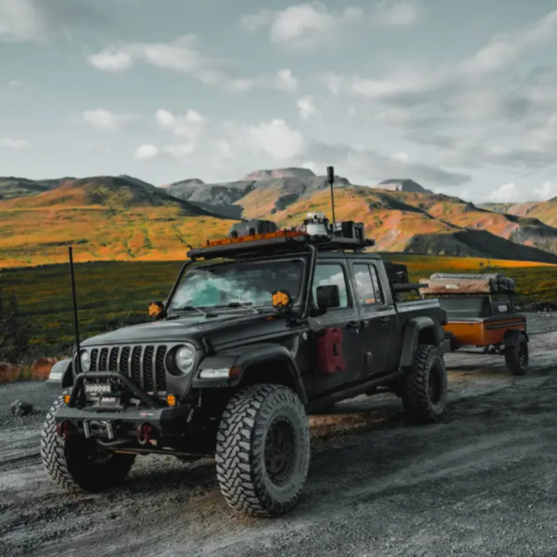 Image of a black Jeep Gladiator overland truck with an extensive roof rack and an antenna, towing a small orange trailer on a dirt road. The vehicle is set against a backdrop of rolling mountains with patches of green and yellow vegetation under a cloudy sky