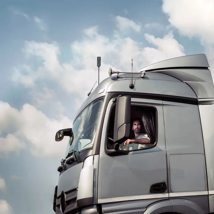 Image of a truck driver looking out of the window of a large silver truck, with a black antenna visible on the roof against a background of a cloudy sky