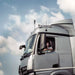 Image of a truck driver looking out of the window of a large silver truck, with a black antenna visible on the roof against a background of a cloudy sky
